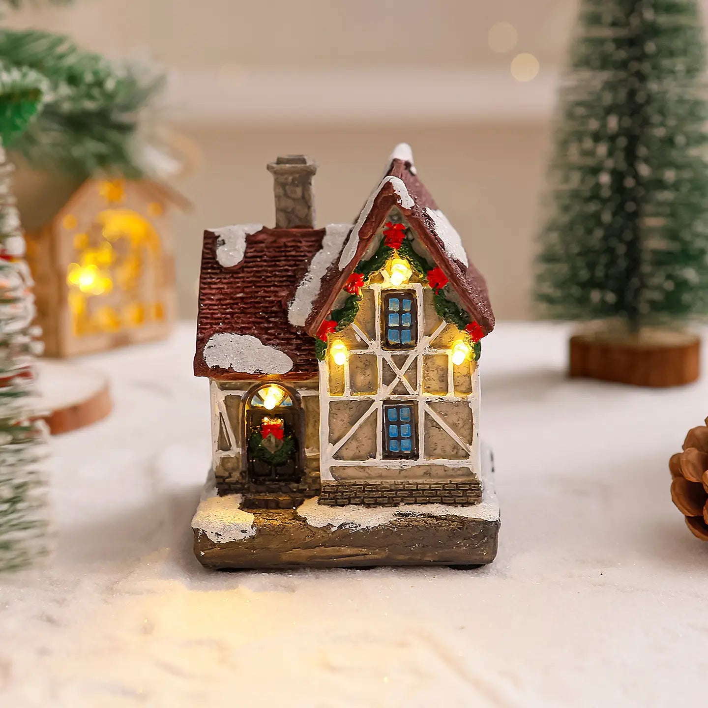 Ornement de maison de village de Noël éclairée présentant une petite chaumière miniature illuminée, un toit brun, des accents blancs comme enneigés, et des guirlandes vertes festives avec des baies rouges.