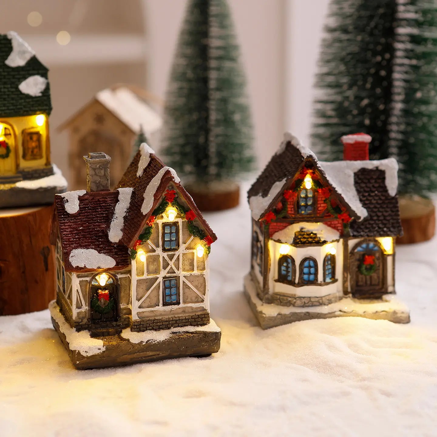 Deux ornements de maison de village de Noël miniature éclairée avec des toits couverts de neige, façades beige et brune à colombages, des lumières chaudes aux fenêtres, et des guirlandes de couronnes festives sur les portes