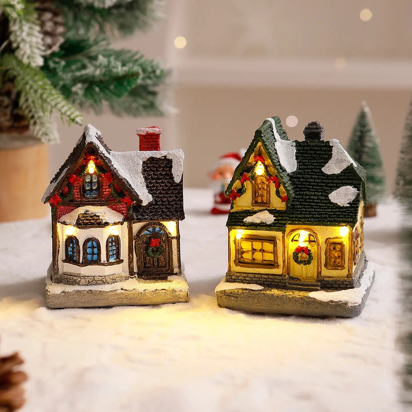Deux ornements de maison de village de Noël miniature éclairée, dont l’un avec un toit rouge et des accents blancs comme enneigés, illuminé chaleureusement, et l’autre avec un toit vert et des couronnes festives doucement illuminées.