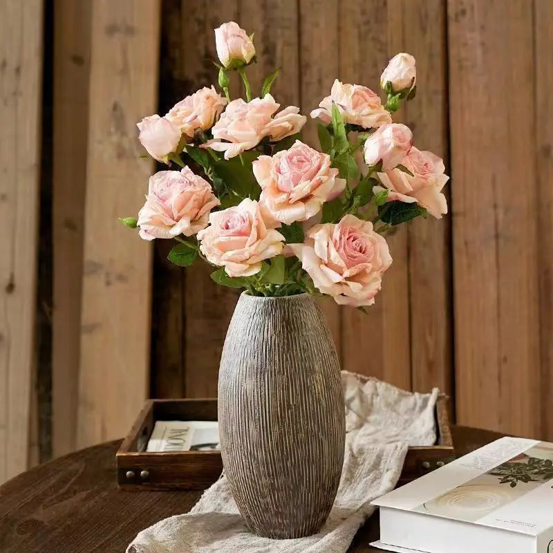 Eine hohe, strukturierte graue Keramikvase hält einen Strauß aus zartrosa verwelkten Stielrand-Rosen in frontaler Ansicht und präsentiert Vintage-Heimdekoration.