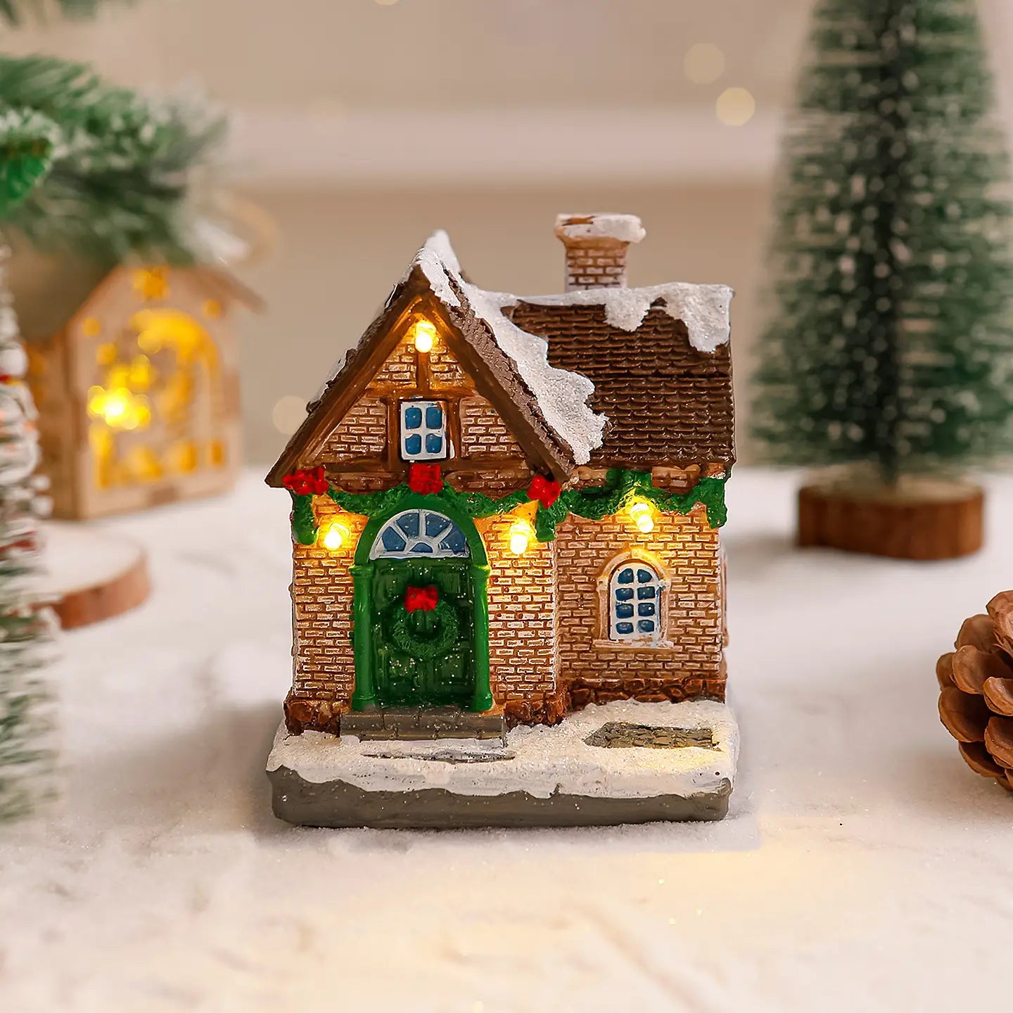 Ornement de maison de village de Noël éclairée présentant une façade miniature en briques brunes, une porte verte et des cadres de fenêtres décorés de baies rouges et de guirlandes de houx, des tuiles de toit couvertes de neige, et des lumières jaunes chaudes.