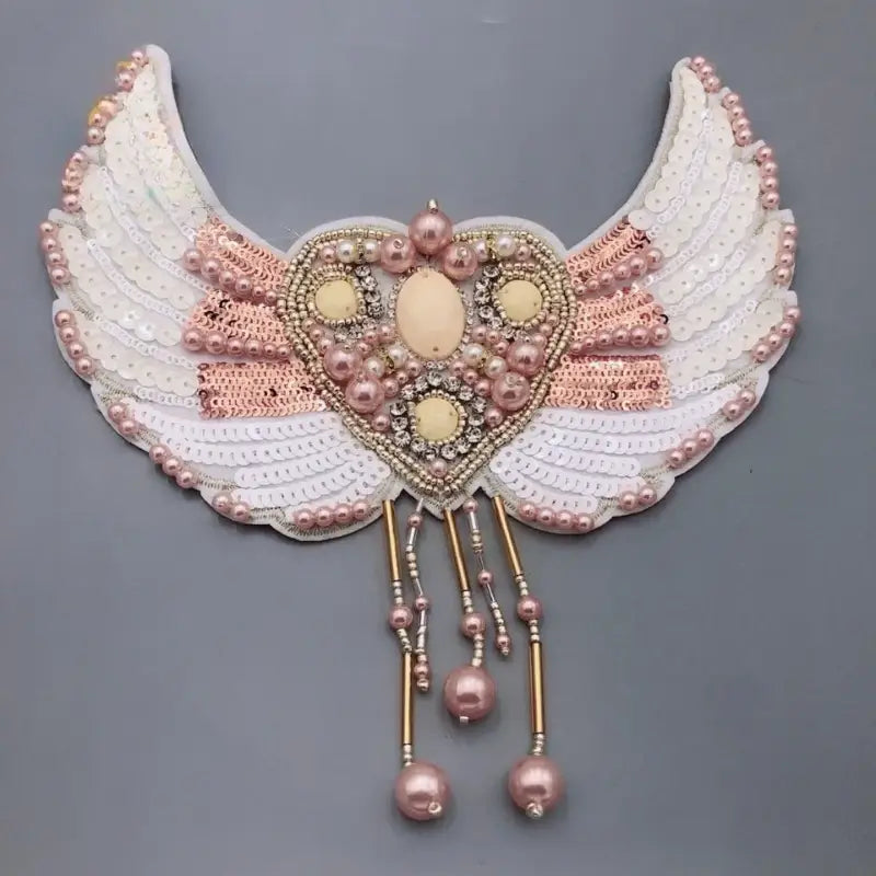 Bordu de perles broche en forme d’aile d’ange pour sac, pièce d’ornement comportant un centre en forme de cœur orné de perles roses et crème, de sequins, de perles, et rehaussé de brins de perles pendants.