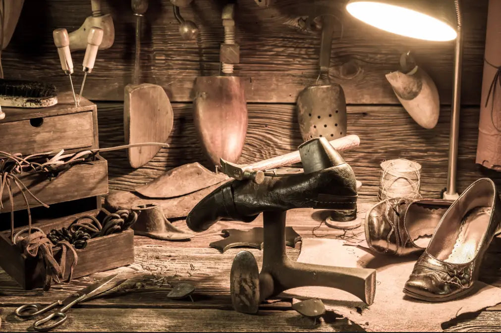 A black leather shoe being repaired with a cobbler’s tool alongside men light blue oxford dress shoes.