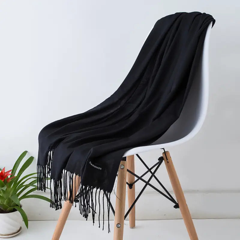 やわらかな質感と繊細なフリンジを備えた黒の女性用カシミヤ風冬用スカーフ／ショールで、モダンな椅子の上にエレガントに掛けられています。