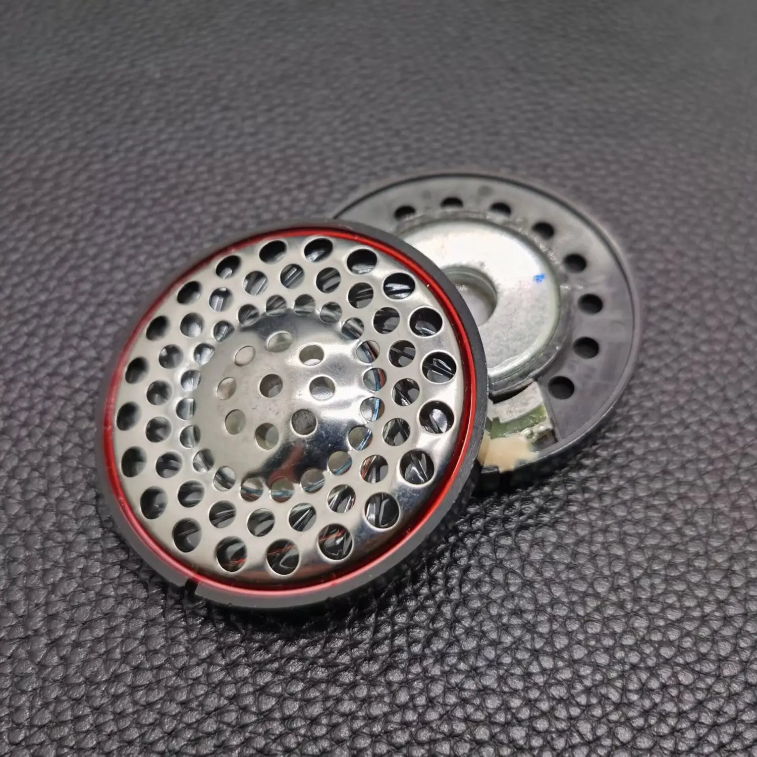 Altavoz de repuesto para auriculares Ath-ad900x que presenta una rejilla circular de metal perforado plateada con anillo de goma roja y carcasa de plástico negro.