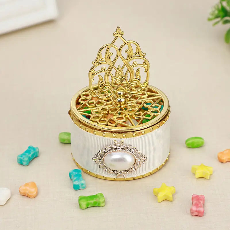 Une boîte à bijoux blanche cannelée avec un couvercle bordé d’or, arborant un design en filigrane orné et une perle centrale accompagnée de cristaux étincelants, utilisée comme boîte à bonbons de mariage dorée décorative, contenant une perle-bonbon en guise de cadeau.