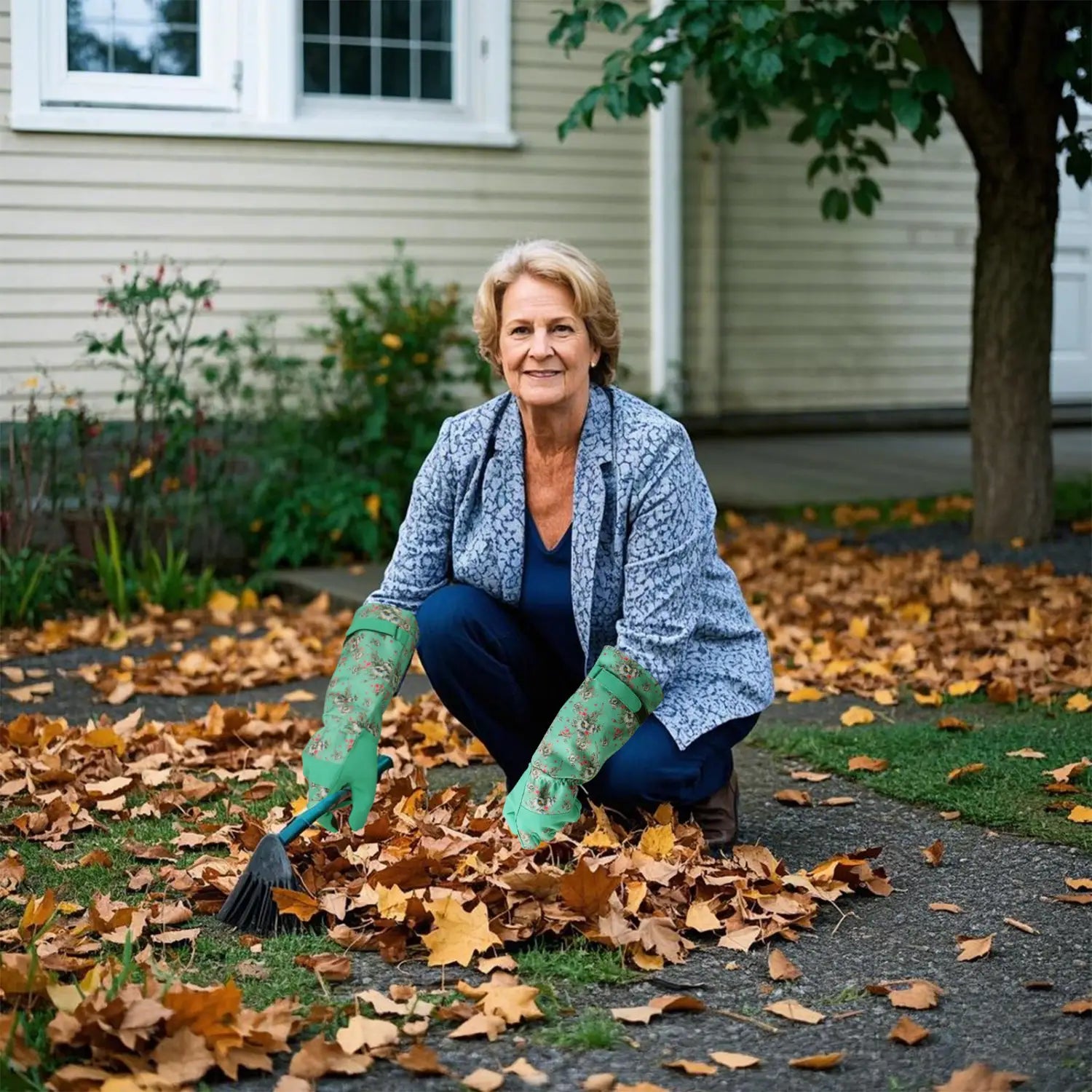 Une femme à l’extérieur portant des gants de jardinage floraux verts respirants et anti-épines pour femmes s’accroupit, tenant un râteau à manche noir au milieu des feuilles d’automne tombées.