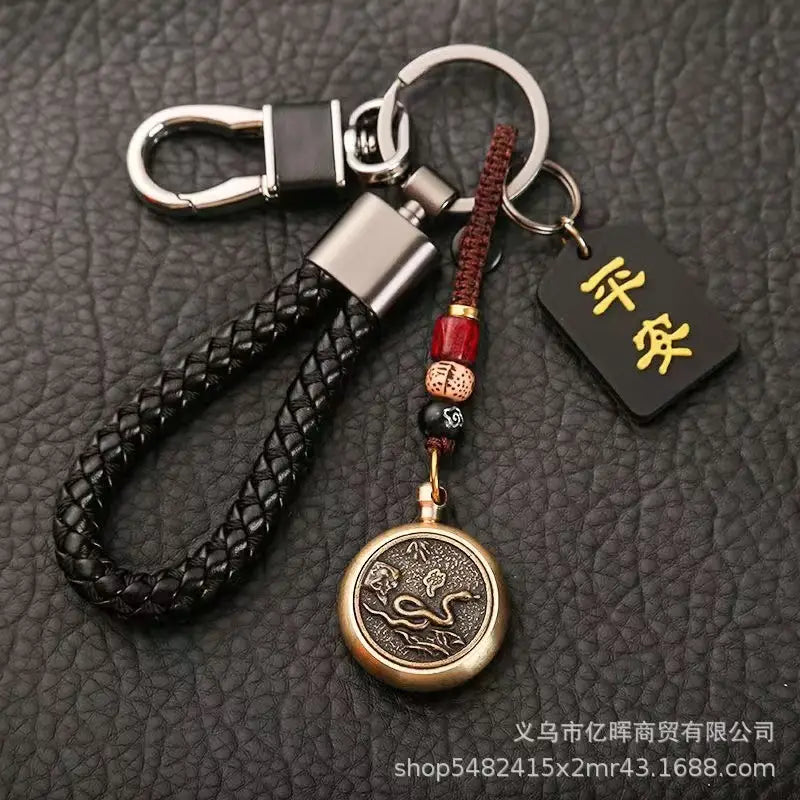 赤と黒のビーズでアクセントを施し、刻印された「蛇」のデザインをあしらったブロンズ円形ペンダントと、金色の文字が記された小さな黒いタグを備えた黒の編みレザー製キーチェーン（中国の干支をモチーフにした編みレザー製ラッキーペンダント）。