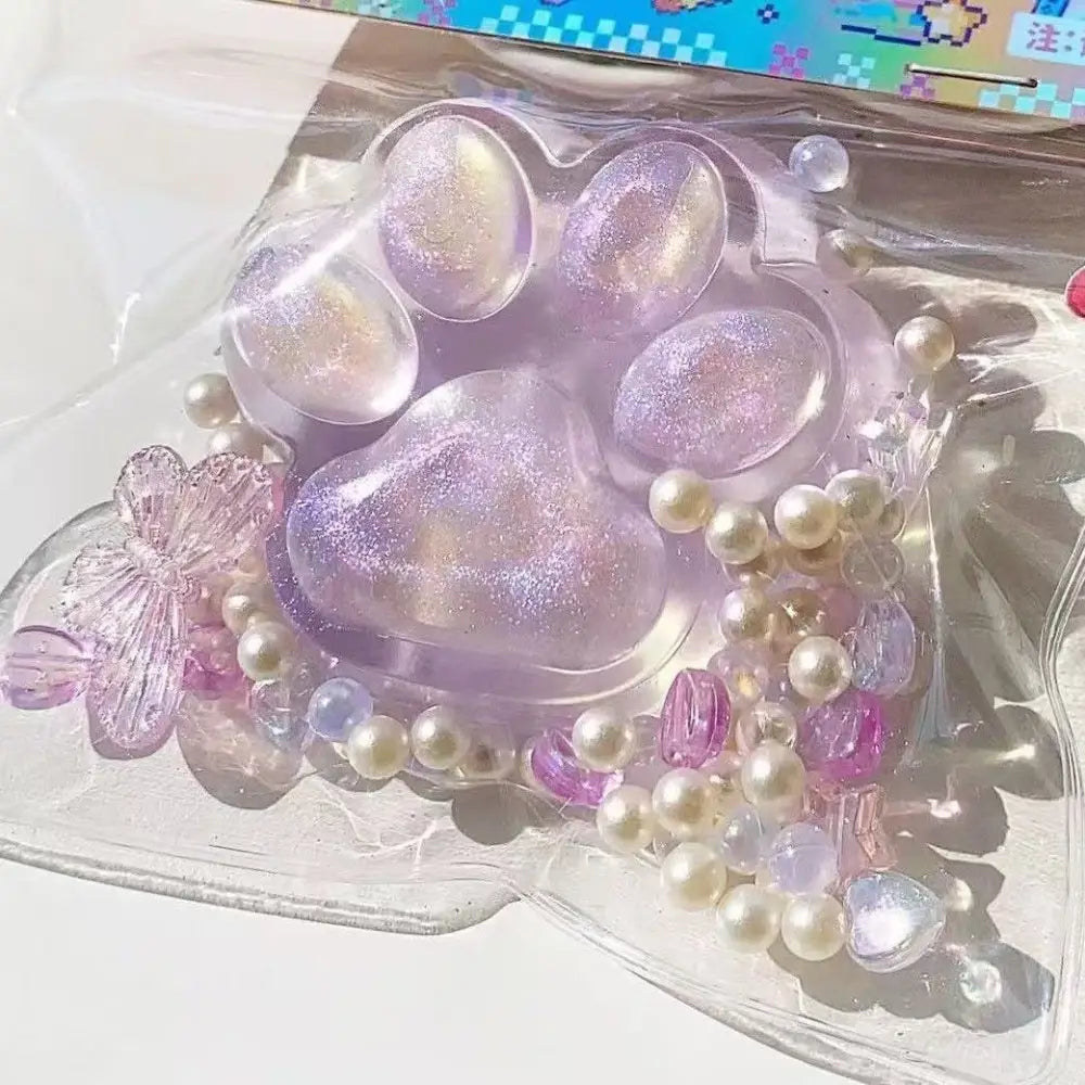 amuleto exprimible de resina translúcida con forma de pata de gato en color lavanda, con destellos de purpurina, rodeado de cuentas perladas y de cristal, para utilizarlo como juguete exprimible de pata de silicona con liberación lenta y gradual, a fin de aliviar el estrés.