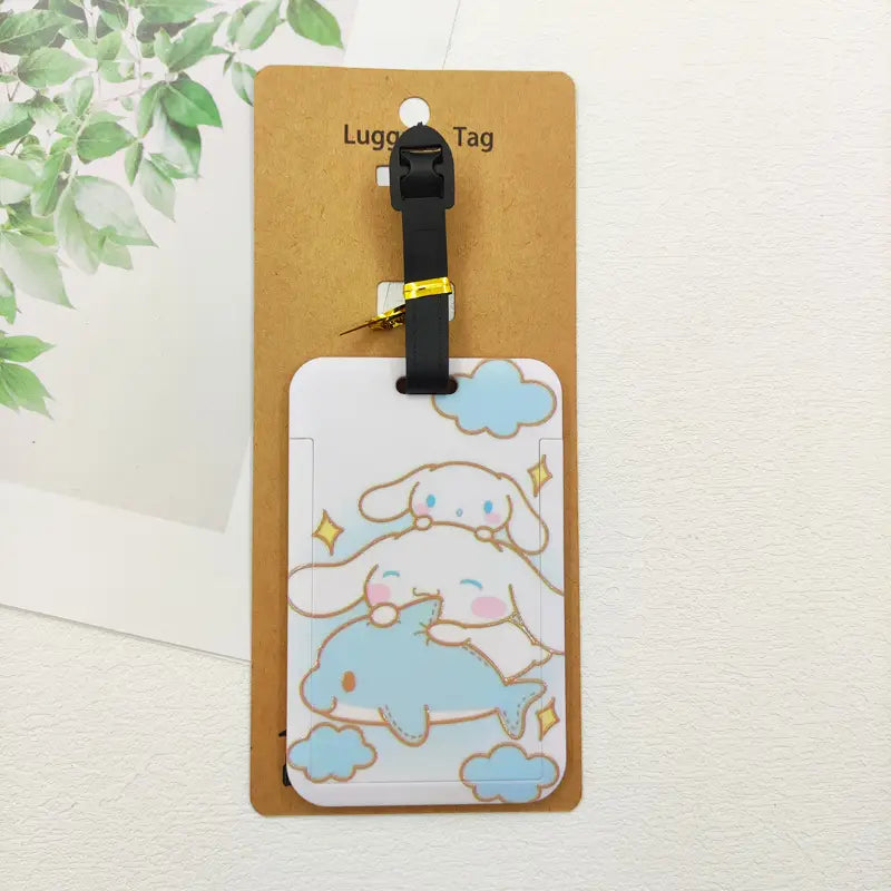 Étiquette de bagage rectangulaire blanche Hello Kitty, avec un mignon design de dessin animé de Cinnamoroll chevauchant un dauphin bleu, des nuages bleu clair et des étoiles jaunes, fixée à une sangle noire pour l’identification de la valise.