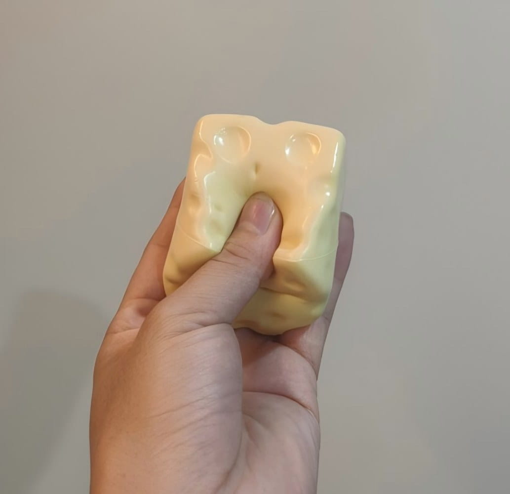 ein blassgelbes, knetbares Sinnes-Plaything, geformt wie ein Käsewürfel mit typischen Löchern, die als Käsewürfel-Stressball dienen.
