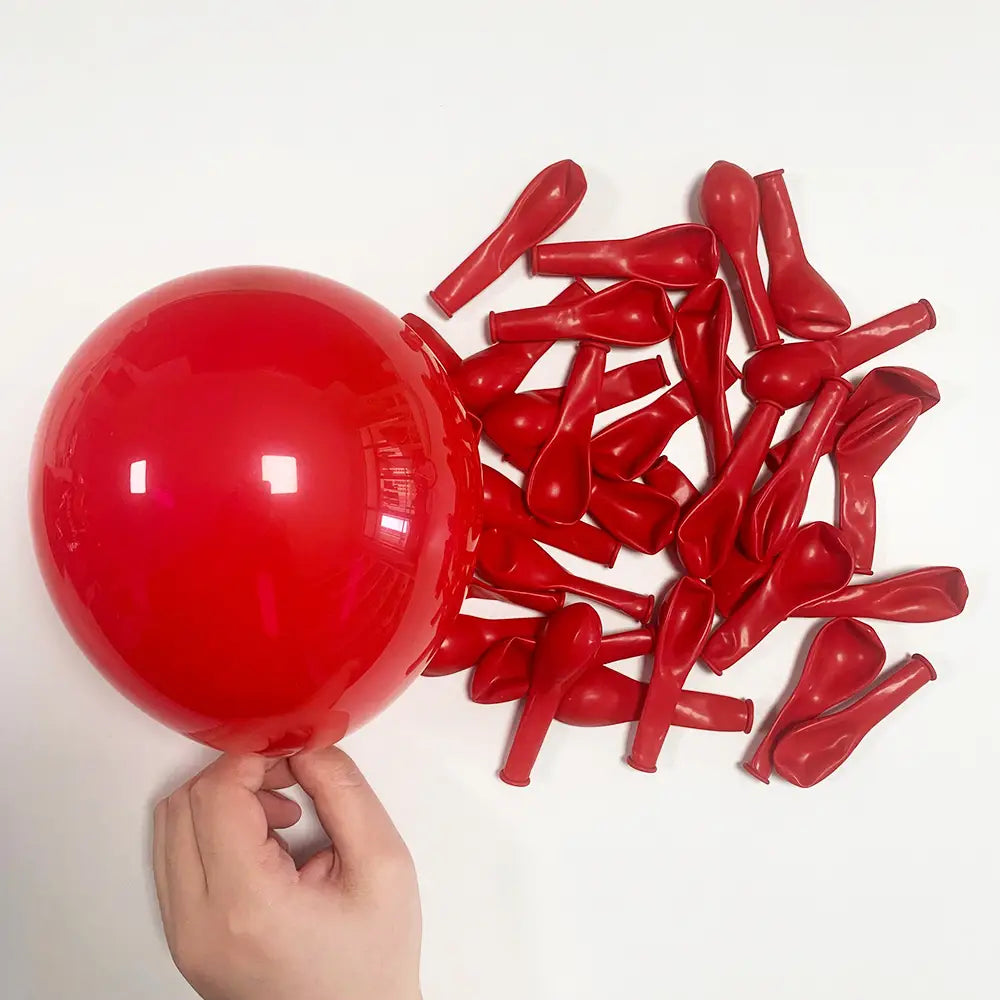 Ein einzelner aufgeblasener roter Latexballon, der von einer Hand gehalten wird, steht im Kontrast zu einer Ansammlung entfalteter roter Latexballons im roten-schwarzen Ballon-Girlandenbogen-Set zur Partydekoration.