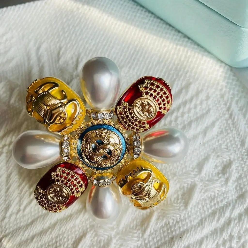 Broche florale de luxe Chanel, présentant des pétales d’émail à ton doré en rouge et jaune, rehaussés de perles blanches et de cristaux scintillants, entourant un emblème central bleu et or, conçue comme une broche de perles designer en forme de coléoptère, épingle sertie de rhinestones.