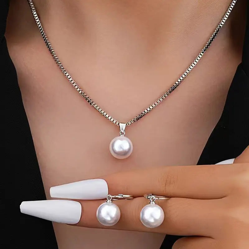 Un ensemble de bijoux en tons argentés, comprenant un collier à pendentif et des boucles d’oreilles assorties à tige, avec une seule perle ronde de rivière.