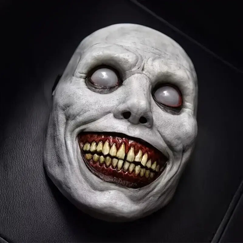 A gray textured latex full face horror mask with exaggerated features including wide hollow eyes one glowing white one red and a grotesquely smiling mouth with yellowed teeth for creepy Halloween costume accessory.
