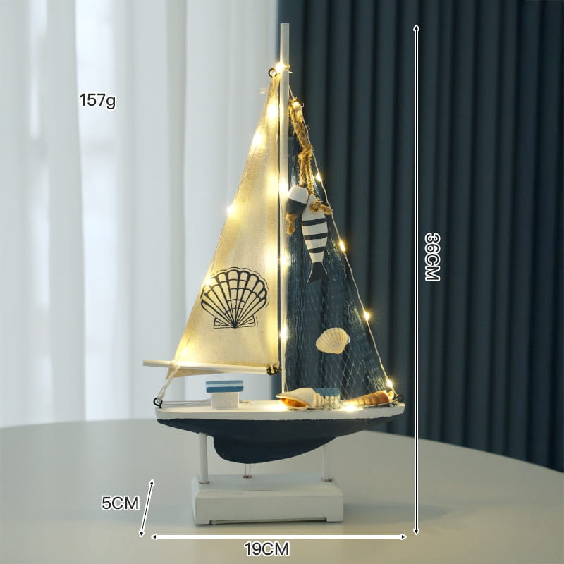 Eine Figurendeko im mediterranen Stil aus Holz mit LED-Licht für ein Segelboot im Wohnbereich: weißer Sockel, dunkelblauer Rumpf, zwei Segel in Cremefarbe mit einem schwarzen Muschel-Emblem, dunkelblau, verziert mit Lichterketten, kleine Muscheln und gestreifte Fische.