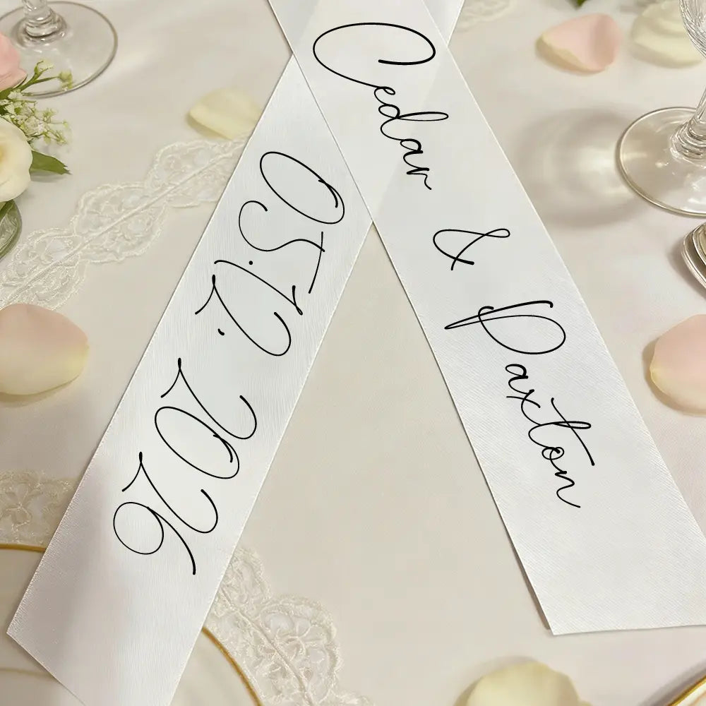 Un ruban de bouquet de mariage en satin blanc personnalisé avec un lettrage cursif noir indiquant caden et daxton 05.27.2026, avec des noms propres et une date.