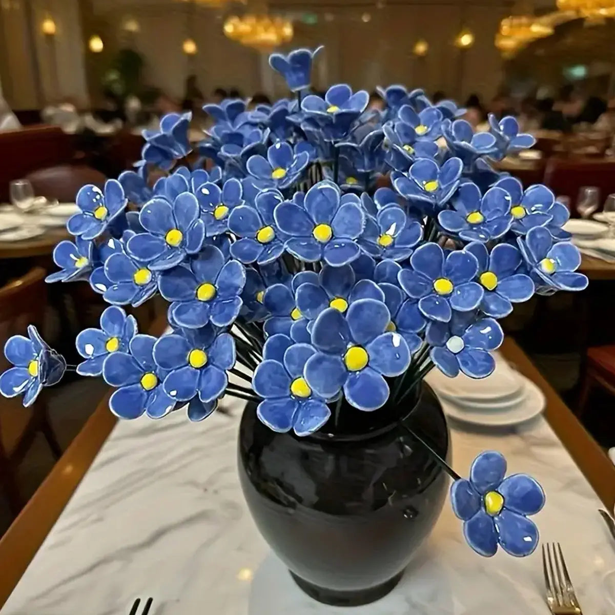 A vibrant blue ceramic artificial flowers arrangement with yellow centers displayed in a sleek black decorating vase.