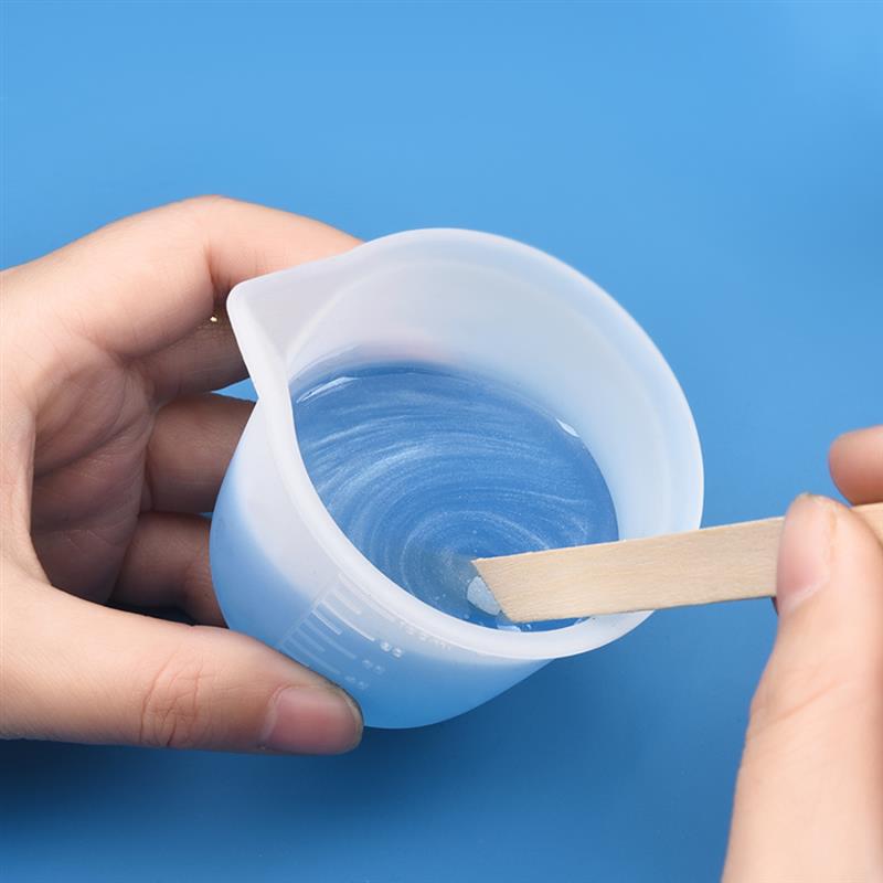 Light blue translucent plastic measuring cup with a spout holding clear liquid being stirred by a wooden stick for epoxy resin mixing tool set for diy jewelry making.