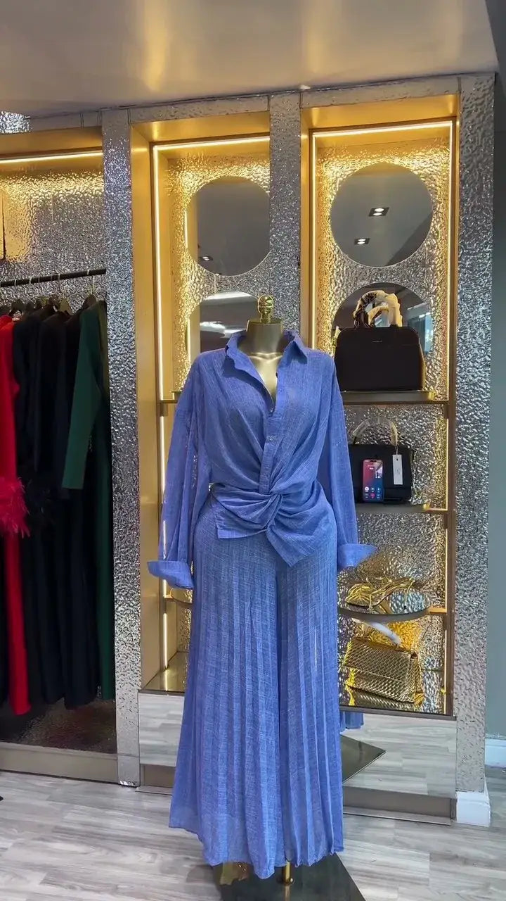 Ein fließendes blaues Falten-Maxikleid, kombiniert mit einem passenden, an der Taille gebundenen Hemd, auf einem Schaufensterkörper in einer Boutique-Umgebung gezeigt, die das Damen-Set mit Faltenrockhose im weitem Bein, bestehend aus einem langärmeligen Outfit, präsentiert