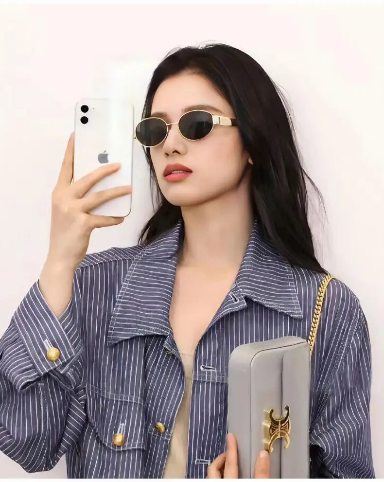 Un bolso de mano rectangular gris con broche metálico en tono dorado y correa de cadena, presentado junto con gafas de sol ovaladas para mujer y hombre con montura dorada.