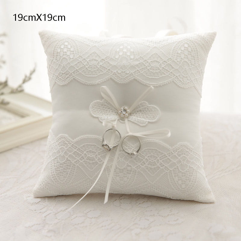 Almohada blanca para anillos de boda de encaje con un delicado lazo floral y dos anillos plateados colocados en su centro para uso como almohadilla portadora de arras.