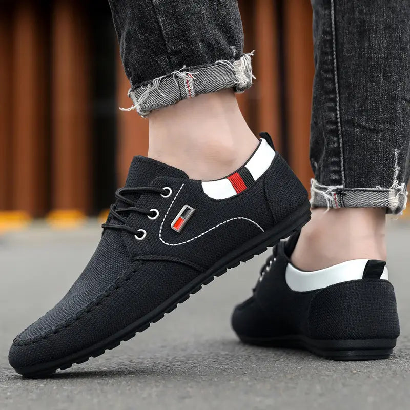 Black canvas lace-up men’s casual sneakers with white and red accent stripes on the heel tab and a stitched logo patch designed for lightweight daily footwear shown from the back side.