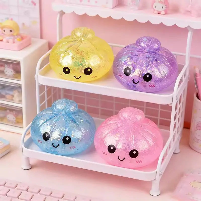cuatro juguetes exprimibles de panecillo al vapor coloridos y con purpurina, con caritas de dibujos animados adorables, exhibidos en una estantería blanca de dos niveles junto con un recipiente para vapor.