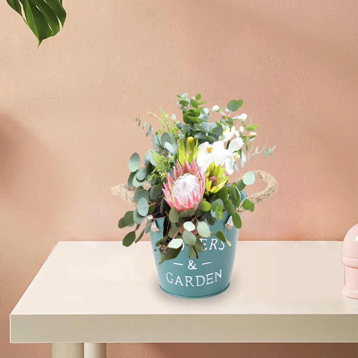 Ein teal-farbener Metall-Blumentopf mit Blumen und im weißen Gartenaufdruck, der eine lebendige Anordnung aus pinken Protea, weißen Orchideen und grünen Eukalyptusblättern hält, als rustikaler Landhaus-Gartenpflanzen-Blumentopf.