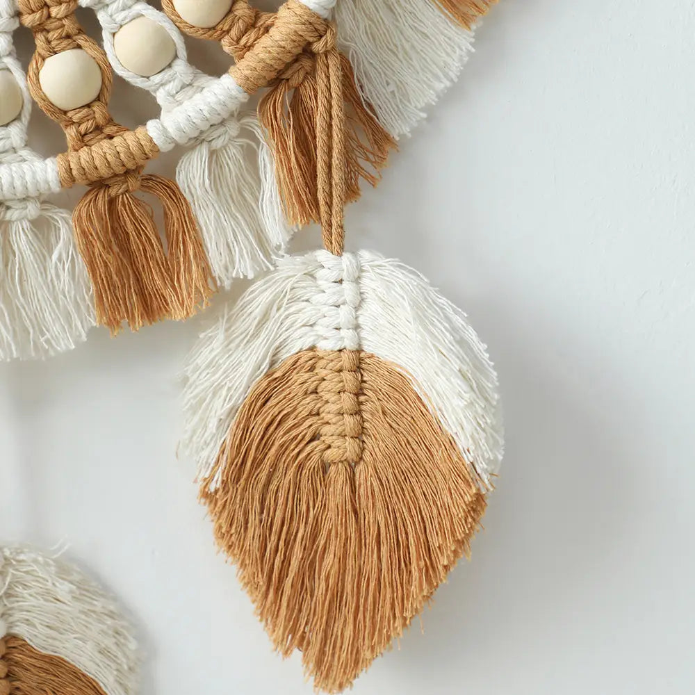 A handcrafted cream and tan macrame feather ornament with intricate woven patterns wooden beads and fringe tassels for boho living room wall mirror home decor.