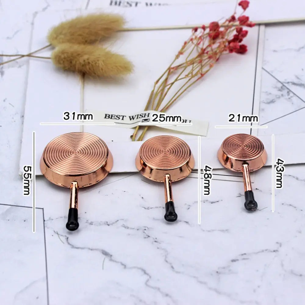 Trois petites poêles à frire de couleur or rose, avec des poignées noires et des surfaces de cuisson circulaires à relief, de tailles variées, provenant d’un ensemble d’ustensiles de cuisine miniature pour maison de poupée.