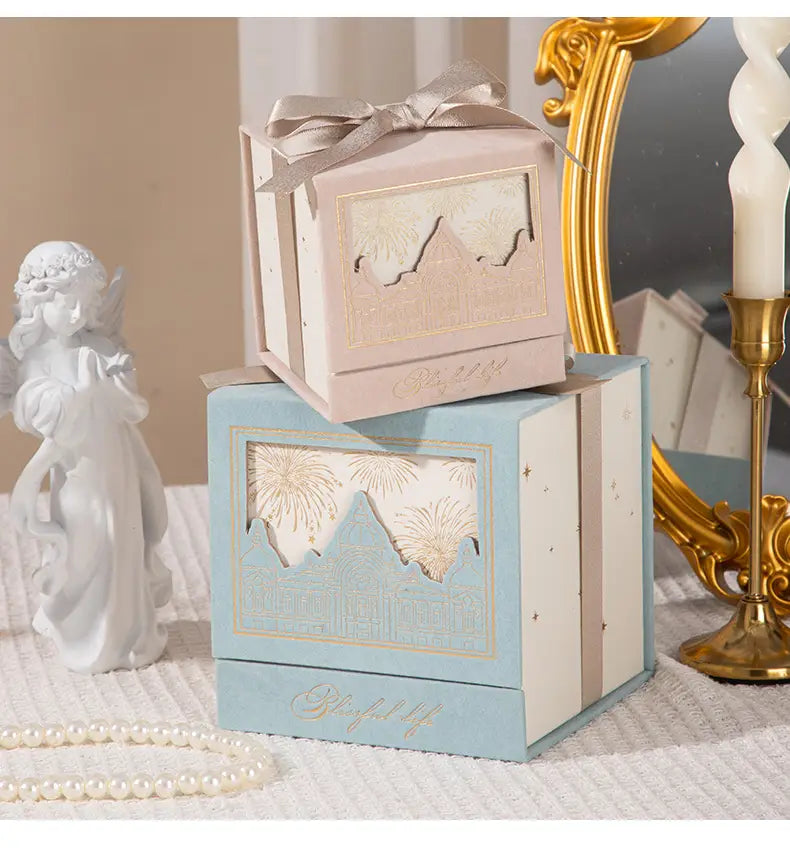 European style wedding gift box candy packaging featuring a pastel blue and cream two-tiered box with gold foil mountain and fireworks design tied with a satin ribbon.