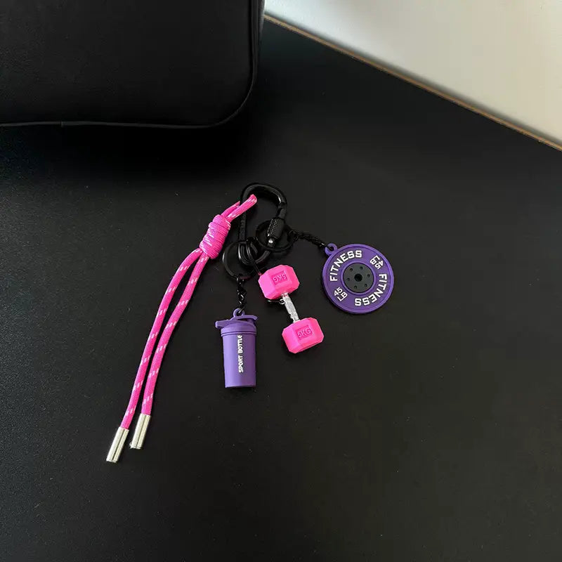 Un porte-clés de fitness rose et violet comprenant une corde tressée avec une breloque de mini-haltère, une breloque de disque de poids et une petite bouteille shaker, pour l’usage avec un sac de sport.