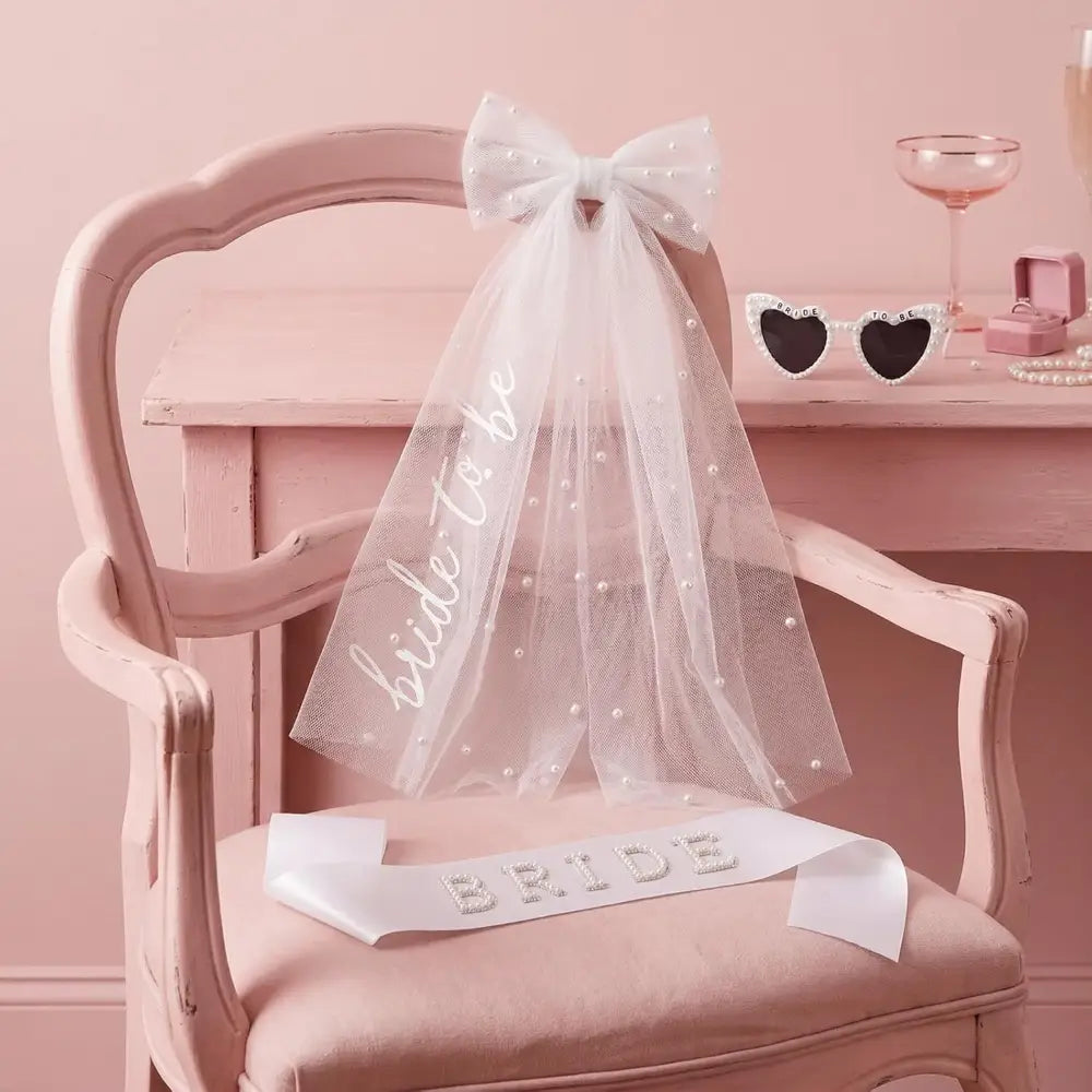 Conjunto de accesorios de la futura novia con un delicado velo de tul blanco adornado con perlas dispersas y un lazo a juego que presenta un texto cursivo de brillo plateado elegante que dice «bride to be».