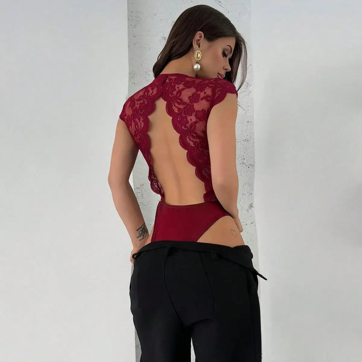 A woman wears a red lace V-neck bodysuit with an open back and scalloped edges paired with black trousers.