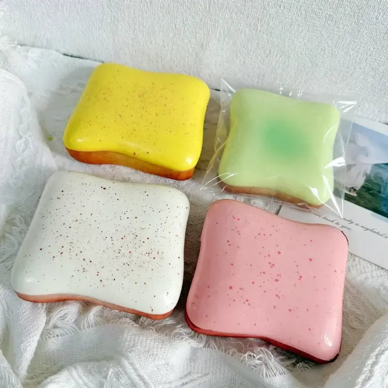 Quatre jouets anti-stress en forme de toast moelleux de forme carrée en silicone, aux couleurs vives, en couleurs macaron : jaune, vert, blanc et rose.