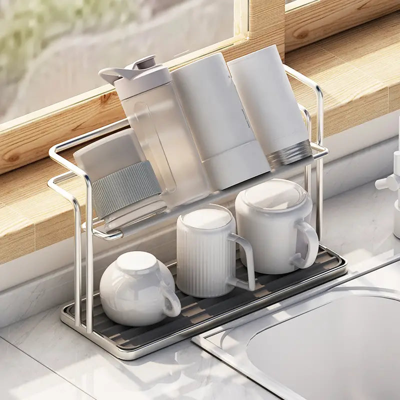 Silver metal stainless steel two-tier kitchen dish rack with a black rubber mat base holding white mugs and containers for mug and cup storage.