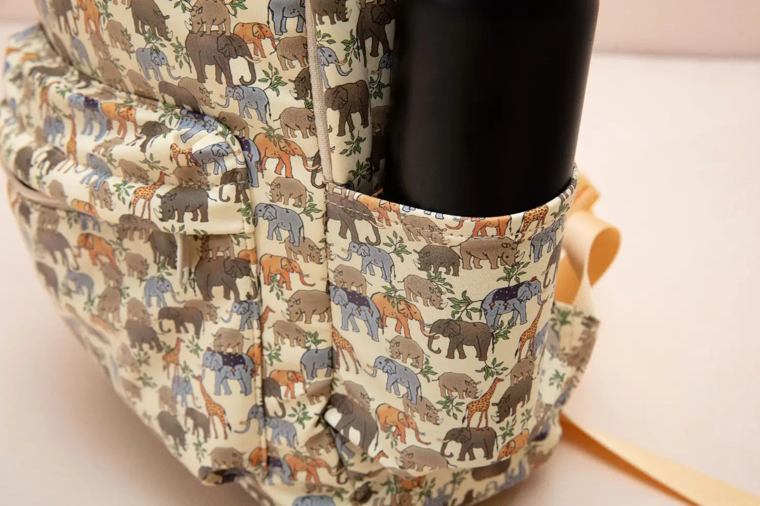 Un sac à dos crème pour enfants en tissu à thème dinosaure, avec un motif ludique d’éléphants, de girafes et d’animaux de safari aux tons terreux atténués, adapté pour l’école.