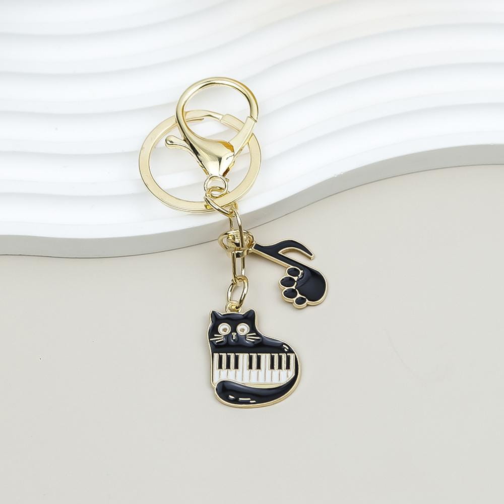 Gold-toned keychain featuring a black enamel cat with piano keys for its body and a matching paw print charm suitable as a music lover gift.