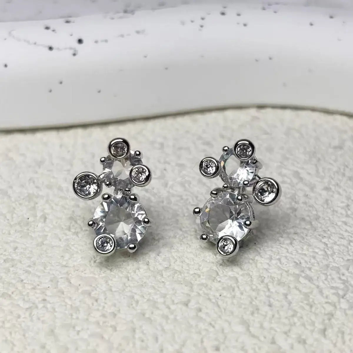 Un par de pendientes tipo botón en tono plateado con cristales facetados transparentes dispuestos en un diseño asimétrico de racimo, de un conjunto de joyería de cristal que incluye un collar de plata, pendientes y una pulsera.