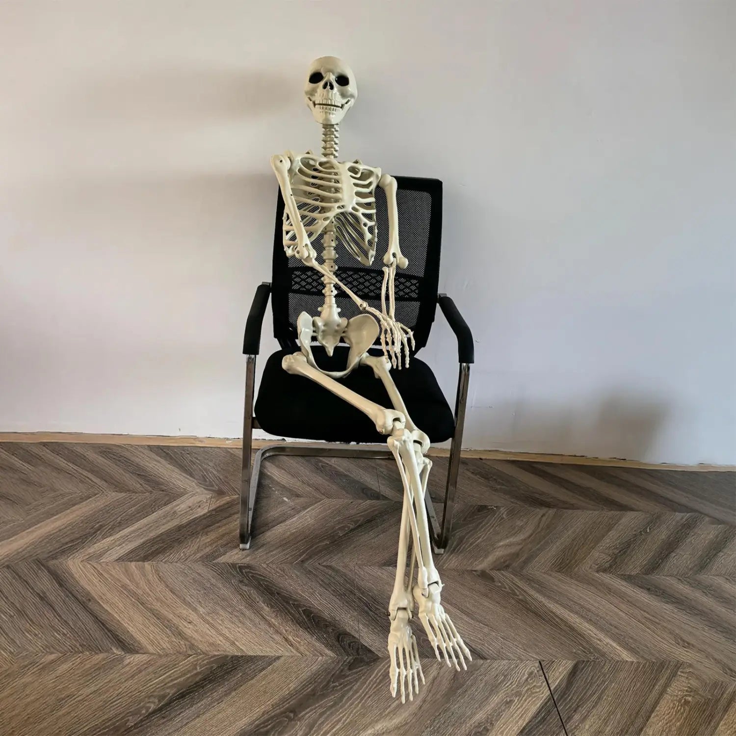 una figura modelo decorativa posable de esqueleto de Halloween de plástico color beige, sentada en una silla de oficina para exhibición de casa encantada.