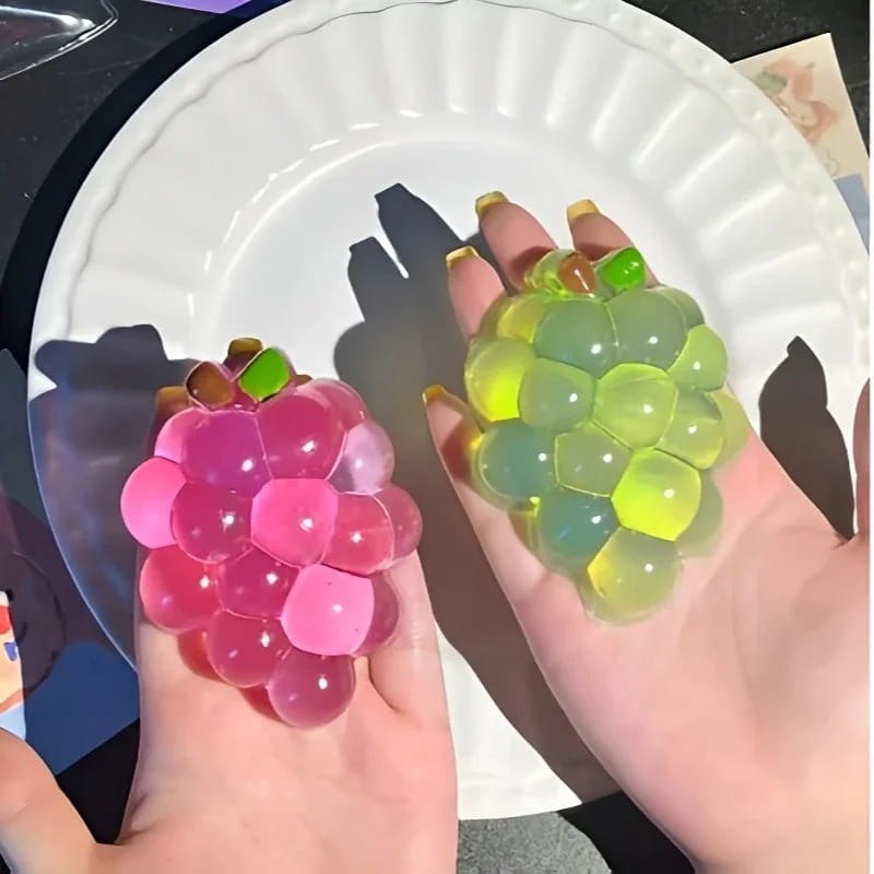 Hände, die rosafarbene und grüne, transparente Trauben-geformte Stressbälle aus dem Taba-Squishy-Grapefruit-Fidget-Spielzeug Taba-licious Fun Fruit-Kollektion halten