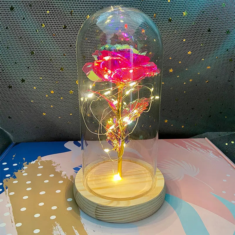 Eine leuchtend pinke und goldene LED-„Eternal Gold Rose“, umschlossen in einer klaren Glaskuppel, mit um den Stiel gewickelten Lichterketten, ruhend auf einem Holzsockel.
