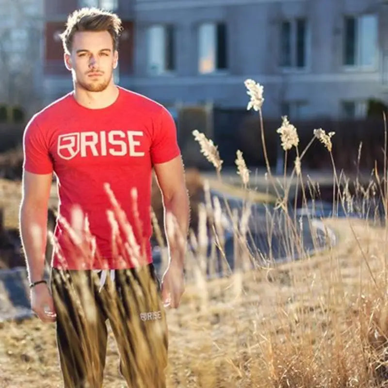 Das Herren-Workout-T-Shirt mit Gold-Rise-Logo ist ein rotes Baumwoll-T-Shirt mit Kurzarm; mit einem weißen Rise-Logo und Wappenemblem, entworfen für den sportlichen Einsatz.