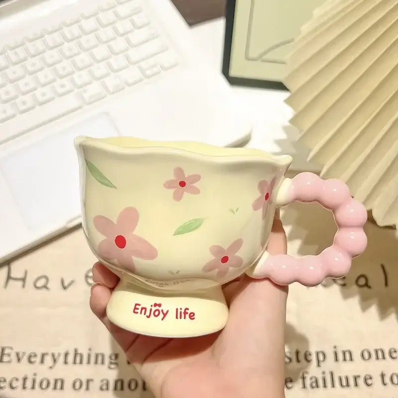 Une tasse tulipe en céramique jaune pastel avec des décors floraux roses et une anse rose perlée, portant la mention « enjoy life » sur sa base, dans une boîte cadeau à motif floral.
