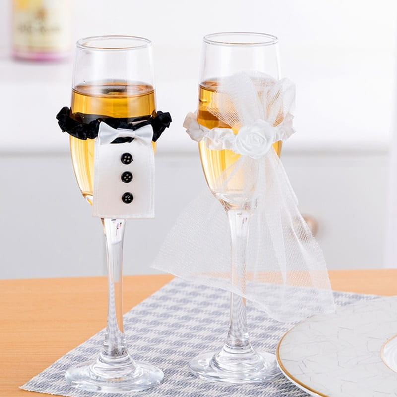 Dos copas tipo champán con cubiertas de copas de vino temáticas de boda que presentan un envoltorio estilo esmoquin negro y blanco y un velo blanco semitransparente en el juego de cubiertas de copas de vino para la novia y el novio.