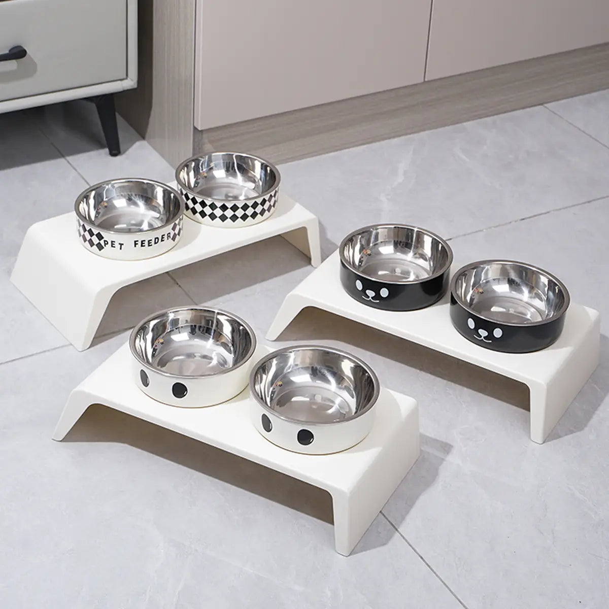 Set of three white plastic elevated dog feeders with raised stands each holding a stainless steel bowl featuring black decorative patterns and cartoon face designs.