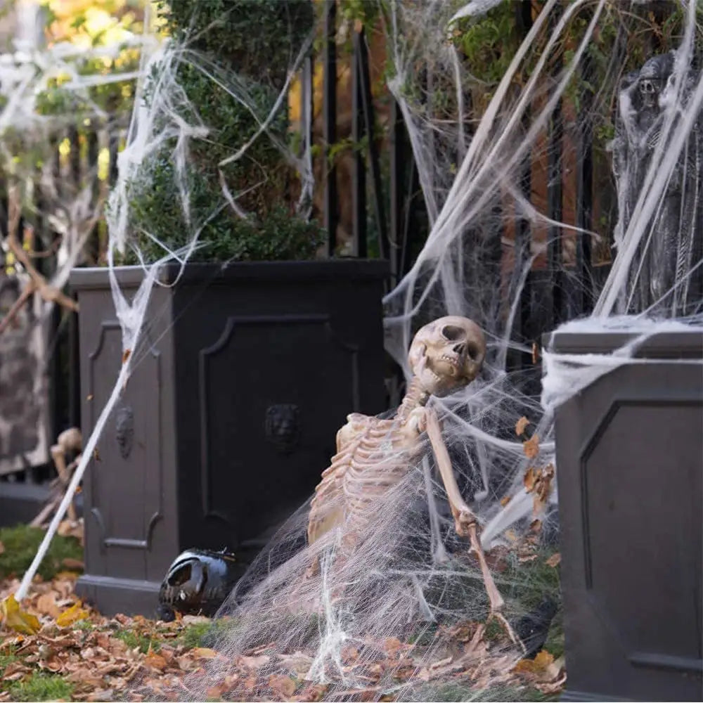 A beige plastic skeleton figure draped in artificial white spiderwebs positioned between dark gray planters as a horror props halloween hanging skull ghost decoration.