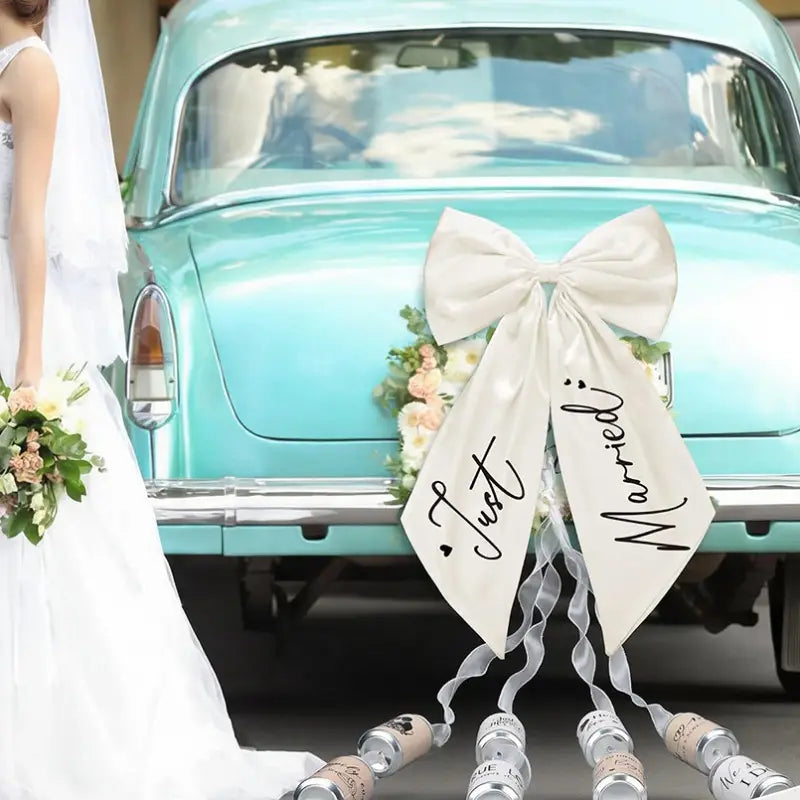Un nœud de voiture « just married » en satin crème, avec une écriture noire cursive, attaché à l’arrière d’une voiture ancienne turquoise pour la décoration de mariage.