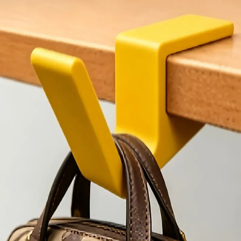 Bright yellow plastic desk edge bag hook holding a brown leather bag strap for backpack storage.